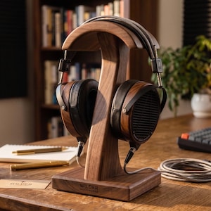 May include: A pair of high-end headphones with wooden earcups and a silver cable rests on a wooden headphone stand. The stand is made of dark wood and has the words "WILDER WOODWORKS" engraved on the base. The headphones are black and brown.