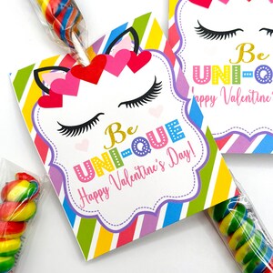 Puede incluir: Una tarjeta de San Valent&iacute;n colorida con tem&aacute;tica de unicornio con el texto "Be Unique! Happy Valentine's Day!" La tarjeta est&aacute; unida a una piruleta con caramelos de colores arco&iacute;ris.