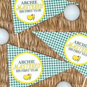 May include: Triangular green and white checkered pennants with the text "ARCHIE MASTERED HIS FIRST YEAR" in a yellow circle, along with a golf course logo. White golf balls are scattered around the pennants. The pennants are on wooden sticks.