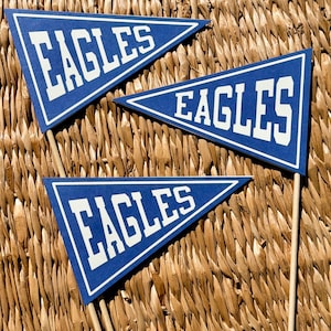 May include: Three triangular blue pennants with white borders and the word EAGLES printed in bold white capital letters. Each pennant is attached to a thin wooden stick, creating a set of decorative sports-themed flags for display.
