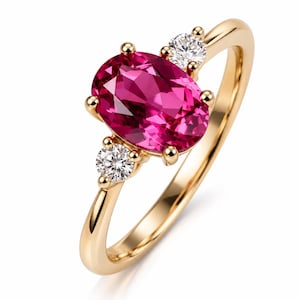 Op de afbeelding: Een gouden ring met een grote, ovaal geslepen, levendig roze edelsteen in het midden. De centrale steen wordt geflankeerd door twee kleinere, rond geslepen, helderwitte edelstenen, één aan elke kant, allemaal vastgezet met gouden klauwen op een gouden band.