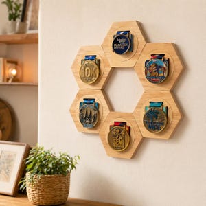Puede incluir: Un expositor hexagonal de madera para medallas con seis medallas. Las medallas redondas tienen cintas azules y varios diseños, incluyendo textos como "Marathon de Paris", "10K" y "Nice". El expositor está montado en una pared clara.