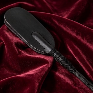 Op de afbeelding: Een zwart lederen peddel met een gladde, afgeronde kop met zichtbare stiksels langs het midden en de randen. Het handvat is omwikkeld met een dun zwart koord, dat overgaat in een lange, gevlochten zwarte koordstaart aan de onderkant.
