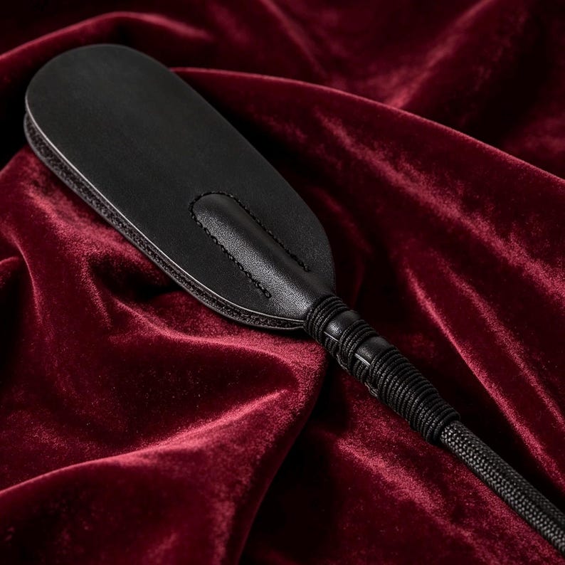 Op de afbeelding: Een zwart lederen peddel met een gladde, afgeronde kop met zichtbare stiksels langs het midden en de randen. Het handvat is omwikkeld met een dun zwart koord, dat overgaat in een lange, gevlochten zwarte koordstaart aan de onderkant.