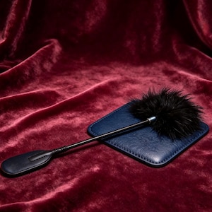 May include: A black feather tickler with a long, slender handle and a flat, rounded leather end, resting on a square, navy blue textured leather pad. The handle features a wrapped grip section and a metallic ferrule connecting to the feathers.