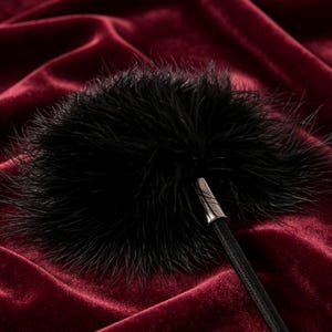 Op de afbeelding: Een zwart haaraccessoire met een verenvormige pompon, bestaande uit een ronde, pluizige tros donkere veren bevestigd aan een dunne, zwarte elastische band met een klein, cilindervormig zilverkleurig metalen verbindingsstuk.
