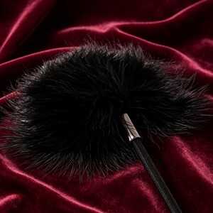 Op de afbeelding: Een zwarte veren pompon bevestigd aan een dunne zwarte elastische band. De pompon is rond en pluizig, met lange, fijne zwarte veren die uitstralen vanaf een centrale zilverkleurige metalen huls die verbonden is met het elastische koord.