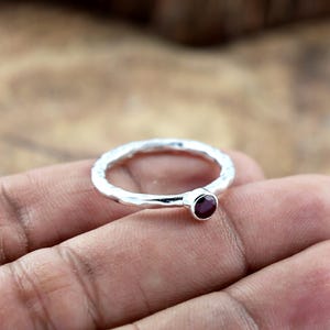 Puede incluir: Un anillo de plata con una textura martillada y una pequeña gema redonda de color rojo oscuro. El anillo está colocado en una mano, mostrando el diseño y el color de la joya. El anillo es una pieza de joyería sencilla y elegante.