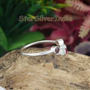 K&ouml;nnte beinhalten: Ein Silberring mit drei kleinen, runden, in Zargenfassung gefassten Edelsteinen in Rot, Wei&szlig; und Dunkelrot. Die Ringschiene ist d&uuml;nn und poliert. Der Text StarSilverIndia ist im oberen Bereich des Bildes &uuml;ber dem Ring zu sehen.