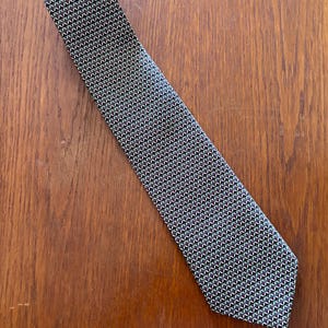 Könnte beinhalten: Eine Krawatte mit einem sich wiederholenden geometrischen Muster aus kleinen Dreiecken in den Farben Schwarz, Weiß und Dunkelrot. Die Krawatte hat ein spitzes Ende und eine konische Form und zeigt ein durchgehendes, dichtes Gitter dieser Formen.