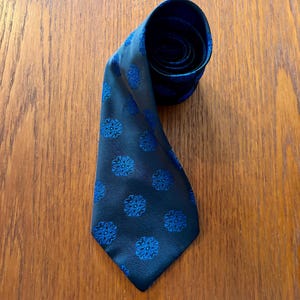 Könnte beinhalten: Eine dunkelblaue Krawatte mit einem sich wiederholenden Muster aus leuchtend königsblauen Blumenmotiven. Die Krawatte ist am oberen Ende teilweise aufgerollt und zeigt eine glatte, leicht reflektierende Stoffstruktur über die gesamte Länge.