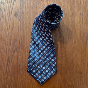 May include: A dark blue necktie featuring a repeating pattern of small white circular symbols connected by thin diagonal red lines. The tie is rolled at the top and displays a pointed end at the bottom, showcasing a consistent geometric design.