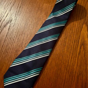 May include: Navy blue tie with alternating stripes of white, teal, and light blue. The tie is diagonally positioned on a wooden surface, showcasing its pattern and design. A classic accessory for formal or business attire.
