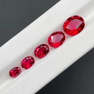 May include: Five oval-cut, vibrant red gemstones of varying sizes are arranged diagonally on a white surface. The gems exhibit a high degree of clarity and brilliance, reflecting light. The largest gem is at the top right, with the smallest at the bottom left.