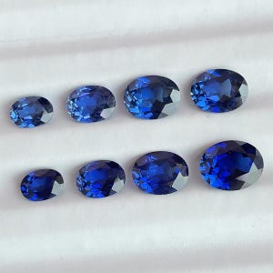 May include: Eight oval-cut, faceted gemstones in varying sizes are arranged on a white surface. The gems display a deep, vibrant blue color, with multiple facets catching the light. The arrangement showcases the gems' brilliance and clarity.