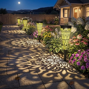 May include: A series of four cone-shaped solar garden lights with intricate cut-out patterns, emitting a warm yellow glow that projects elaborate, symmetrical floral-inspired shadows onto a stone walkway lined with colorful flowers and greenery.
