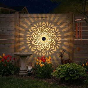 May include: A wooden fence illuminated by a solar-powered light projecting a mandala pattern. The garden scene includes a bird bath, gnome statue, deer statue, wind chimes, and colorful tulips. The night sky is visible in the background.