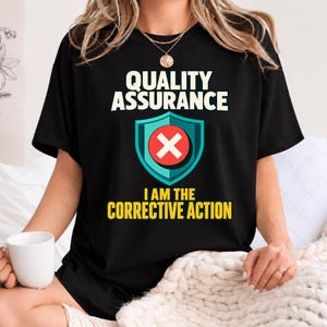 May include: A black short-sleeve t-shirt featuring the white text "QUALITY ASSURANCE" above a teal shield icon containing a red circle with a white "X". Below the shield, the yellow text reads "I AM THE CORRECTIVE ACTION".