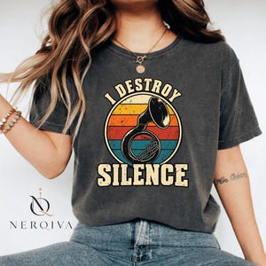 Może przedstawiać: Ciemnoszary t-shirt z krótkim rękawem z nadrukiem graficznym przedstawiającym tubę wewnątrz okrągłego wzoru zachodzącego słońca z poziomymi pasami w kolorze żółtym, pomarańczowym i turkusowym. Napis "I DESTROY SILENCE" jest wydrukowany pogrubionymi, kremowymi literami.