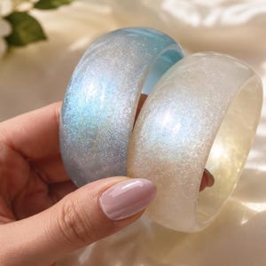 May include: Two thick, rounded bangle bracelets with a smooth, glossy finish. One bracelet is a translucent light blue, and the other is a translucent off-white. Both bangles feature a fine, shimmering glitter embedded throughout the material.