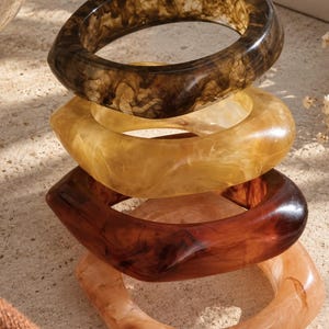 Può includere: Una pila di quattro braccialetti rigidi in resina con una finitura liscia e lucida. I bracciali presentano motivi marmorizzati in tonalità marrone scuro, ambra traslucido, mogano intenso e pesca cremoso, con un effetto vorticoso unico.
