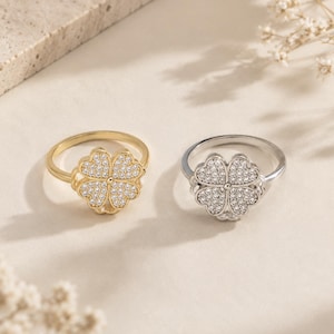 Op de afbeelding: Twee ringen met klaverbladontwerpen. Eén ring is goudkleurig, de andere zilverkleurig. Elke klaver is bezet met kleine, heldere edelstenen. De ringen worden gepresenteerd op een licht oppervlak.