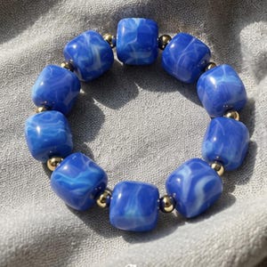 May include: A bracelet featuring ten square, marbled blue and white beads alternating with small, round, metallic gold-tone spacer beads. The word "cornflower" is printed in white text at the bottom of the image, centred below the circular bracelet.