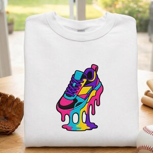 May include: White sweatshirt with a vibrant graphic of a sneaker with a melting effect. The sneaker design incorporates pink, black, blue, yellow, and purple hues. The sweatshirt is made of a soft material, suitable for casual wear.