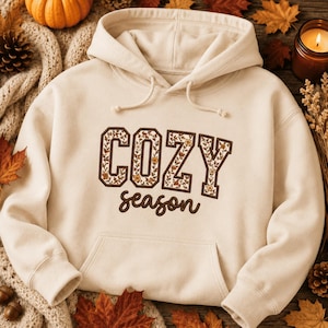 Puede incluir: Una sudadera con capucha de color crema que presenta el texto COZY en letras grandes con borde marrón y relleno de estampado floral, seguido de la palabra season en letra cursiva marrón. La sudadera incluye un bolsillo tipo canguro.