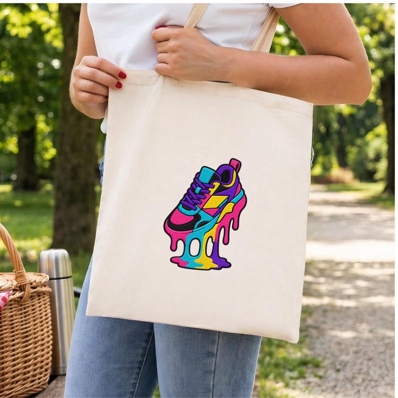 May include: A cream-coloured tote bag with a vibrant graphic of a sneaker with a melting effect. The sneaker design incorporates pink, blue, yellow, and black. The bag is held by a person wearing a white top and blue jeans.