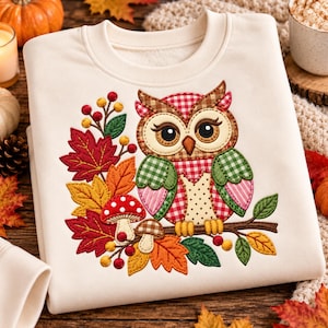 May include: A cream-colored crewneck sweatshirt featuring an embroidered applique design of an owl with a red and white gingham pattern, perched on a branch with colorful autumn leaves, red berries, and two spotted mushrooms.
