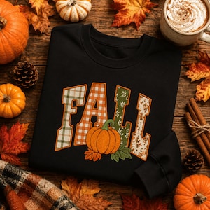 May include: A black long-sleeved sweatshirt featuring the word FALL in large block letters. Each letter has a unique pattern, including plaid, gingham, and floral prints. An embroidered orange pumpkin with green leaves sits below the text.