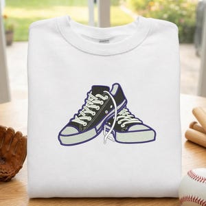 May include: White sweatshirt featuring an embroidered design of black and white sneakers with purple accents. The sneakers are laced up and appear to be a classic style. The sweatshirt is folded neatly on a wooden surface.