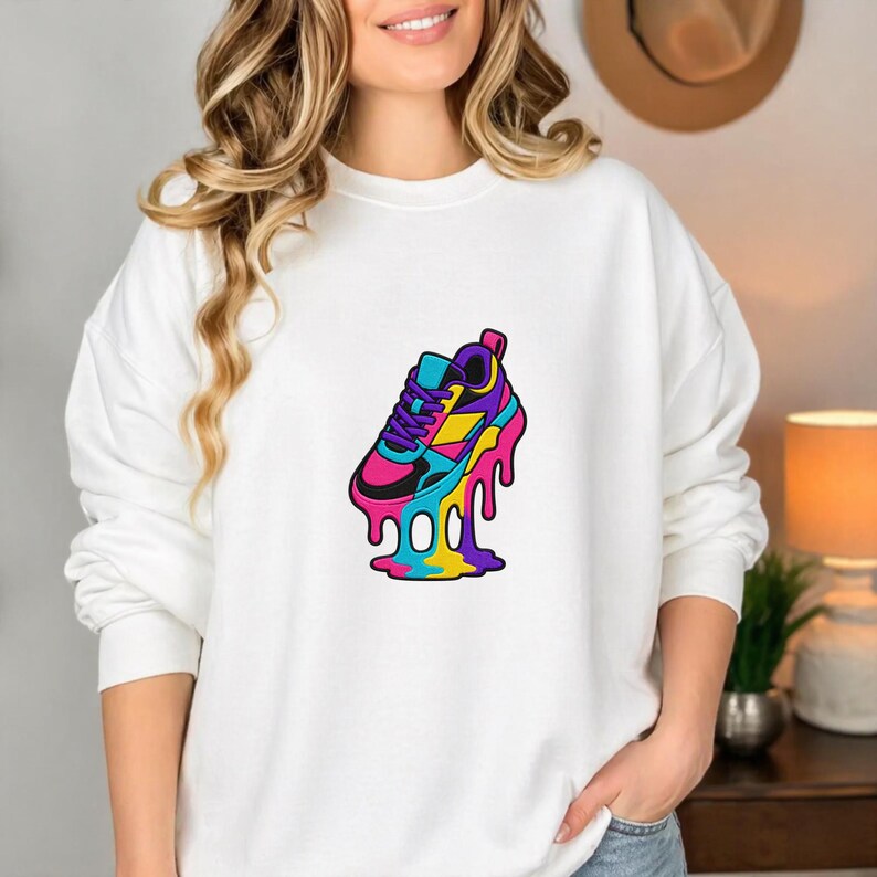 May include: White sweatshirt with a vibrant graphic of a melting trainer. The trainer design features pink, blue, yellow, and purple hues, with black detailing. The graphic has a dripping effect.