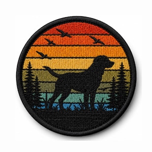 Könnte beinhalten: Runder, gestickter Aufnäher mit einer schwarzen Hundesilhouette vor einem Sonnenuntergang in Orange, Gelb und Blau. Das Design umfasst fliegende Vögel und Tannen. Der Aufnäher hat einen schwarzen Rand.