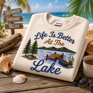 Puede incluir: Una sudadera de cuello redondo color crema con un gráfico bordado de un paisaje lacustre con pinos, un muelle de madera y un pequeño bote. El texto "Life Is Better At The Lake" está cosido con hilo azul y marrón en la parte frontal.
