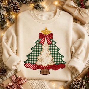 May include: A cream-colored crewneck sweatshirt featuring a central graphic of a stylized Christmas tree. The tree has green and white gingham sections, a white center with gold snowflake patterns, a red polka-dot base, and a red bow.
