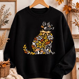 Puede incluir: Sudadera negra con un diseño de gato bordado. El gato está decorado con patrones florales y de hojas amarillas, naranjas y blancas, con una pajarita dorada. El diseño tiene un tema otoñal.