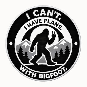 Può includere: Una toppa rotonda in bianco e nero con una silhouette di Bigfoot che fa il segno della pace. Il testo recita "I CAN'T. I HAVE PLANS. WITH BIGFOOT." Montagne e alberi sullo sfondo.