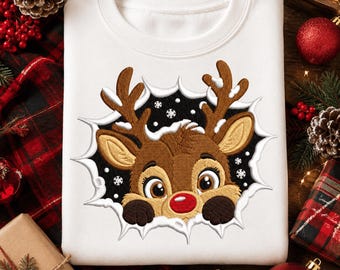 May include: A white crewneck sweatshirt featuring an embroidered graphic of a brown reindeer with a bright red nose. The reindeer appears to be peeking through a jagged, torn-paper style hole against a black background with small white snowflakes.