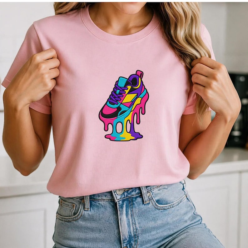 May include: A pale pink t-shirt with a vibrant graphic of a melting sneaker. The sneaker design incorporates blue, pink, yellow, and purple hues. The t-shirt is made of a soft fabric and has short sleeves.