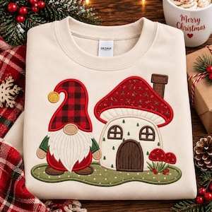May include: A cream-colored crewneck sweatshirt featuring an embroidered design of a gnome wearing a red and black buffalo plaid hat standing next to a mushroom-shaped house with a red roof and a brown door on a green patch of ground.