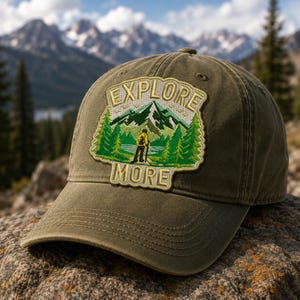 Puede incluir: Gorra de béisbol verde oliva con un parche que muestra un paisaje de montaña y las palabras "EXPLORE MORE". El parche incluye árboles verdes, una figura amarilla y una cordillera. La gorra está hecha de tela resistente y tiene una visera curva.
