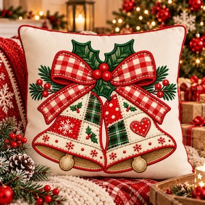 Peut inclure: Un coussin carré blanc cassé avec une broderie représentant deux cloches attachées par un nœud à carreaux rouges et blancs. Les cloches sont ornées de motifs à carreaux verts, de pois rouges, de flocons de neige et d'un cœur. Des feuilles de houx et des baies rouges décorent le haut.