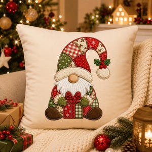 May include: A square cream-colored throw pillow featuring a central embroidered gnome design. The gnome wears a patchwork hat in red, green, and white patterns, has a white beard, and holds a red and green gift box with a bow.