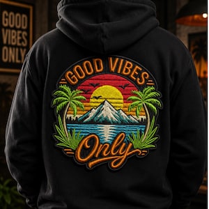 May include: Black hoodie with a colorful embroidered design on the back. The design features a sunset over a mountain and ocean scene, with palm trees and the words "GOOD VIBES ONLY" in a circular arrangement.