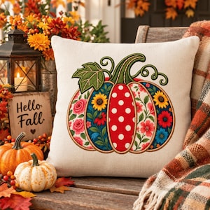 May include: A square off-white throw pillow featuring a central embroidered pumpkin design. The pumpkin is divided into vertical segments with alternating patterns of red with white polka dots, and blue with colorful floral prints of sunflowers.