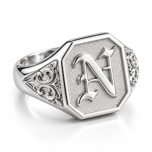 Può includere: Un anello sigillo color argento con una faccia ottagonale dallo sfondo testurizzato e una lettera N in rilievo in carattere gotico. I lati dell'anello presentano complessi intagli a volute che si estendono dalla parte centrale.