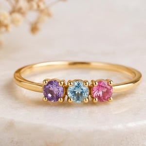 May include: A gold ring featuring three round-cut gemstones set in a row. The stones consist of a purple amethyst on the left, a light blue aquamarine or topaz in the center, and a pink tourmaline or sapphire on the right, all in gold prongs.