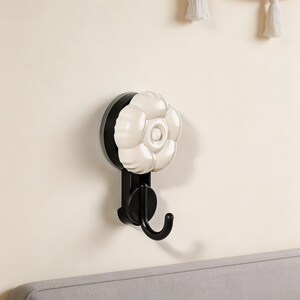 Puede incluir: Un gancho de pared decorativo con un diseño de flor blanca y un gancho negro. El gancho está montado en una pared beige, con un marco de cama gris debajo. La flor tiene un diseño de pétalos detallado y en capas.
